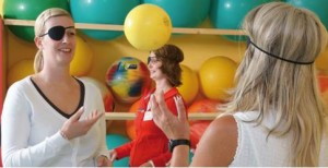 Personen mit Augenklappen machen Übungen mit Bällen in einem Fitnessraum voller bunter Bälle.