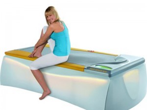 Eine Frau sitzt lächelnd auf einem modernen Massagebett in heller Umgebung.