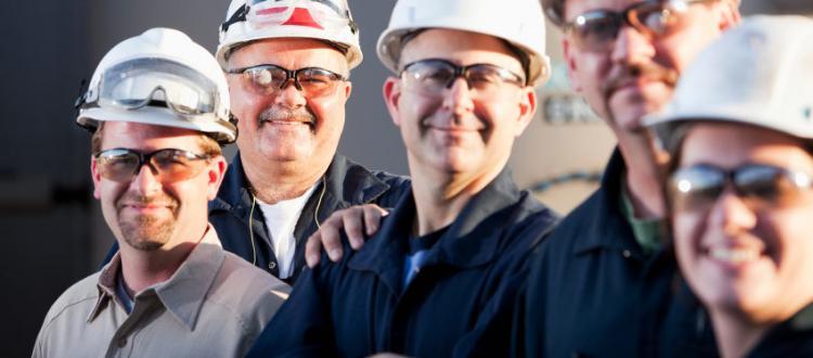Arbeiter stehen nebeneinander, lächeln, tragen Helme und Schutzbrillen, befinden sich auf Baustelle.