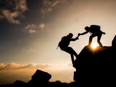 Zwei Menschen, Silhouetten, helfen sich beim Klettern auf einem sonnigen Berggipfel.